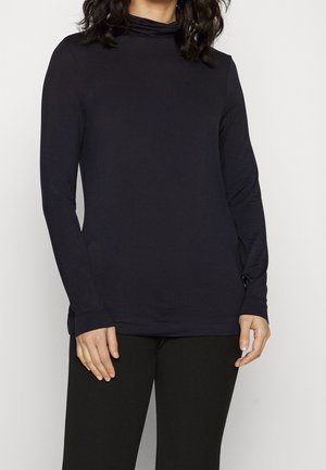 Femme portant un haut à col roulé noir moulant à manches longues et un pantalon noir, debout devant un fond blanc uni.