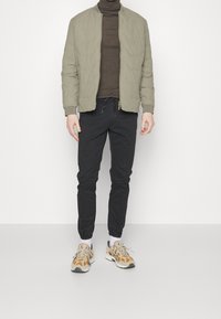 Quiltet olivengrøn bomberjakke, brun rullekrave, sorte koniske bukser og beige sneakers med orange accenter, sat mod en hvid baggrund.