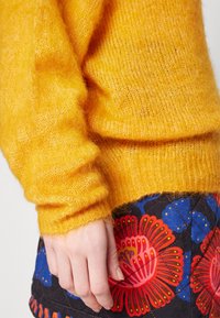 Gul mohairtröja med en mjuk, fluffig textur; åtsittande ärmar. Matchas med en svart kjol med stora blommiga mönster i rött och blått.