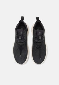 Pantofi sport negri cu un material texturat, șireturi elastice și o talpă albă cu amortizare. Au mânere pe călcâi pentru a fi ușor de încălțat.