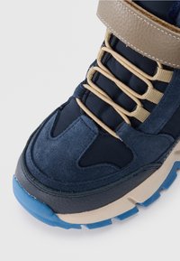 Mörkblå mocka och textil sneakers med ett beige band, elastiska snören och en strukturerad gummisula med blå accenter.