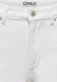 Valge teksade vöökoha lähivaade, millel on hõbedane nööp ja kaubamärgiga silt "ONLY" luku kohal.