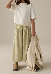 Un t-shirt court beige clair associé à une jupe longue rayée vert et crème. Une veste crème est tenue dans une main. Des chaussures brunes complètent le look.