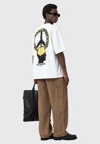 Hvid oversized t-shirt med et sort og gult grafisk motiv på ryggen. Kombineret med beige corduroy-bukser og en sort tote bag.