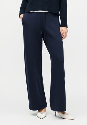 Person iført marineblå bukser med vidde og snøre i taljen, en sort sweater og hvide spidse hæle med blonderstrømper, stående med hænderne i lommerne.