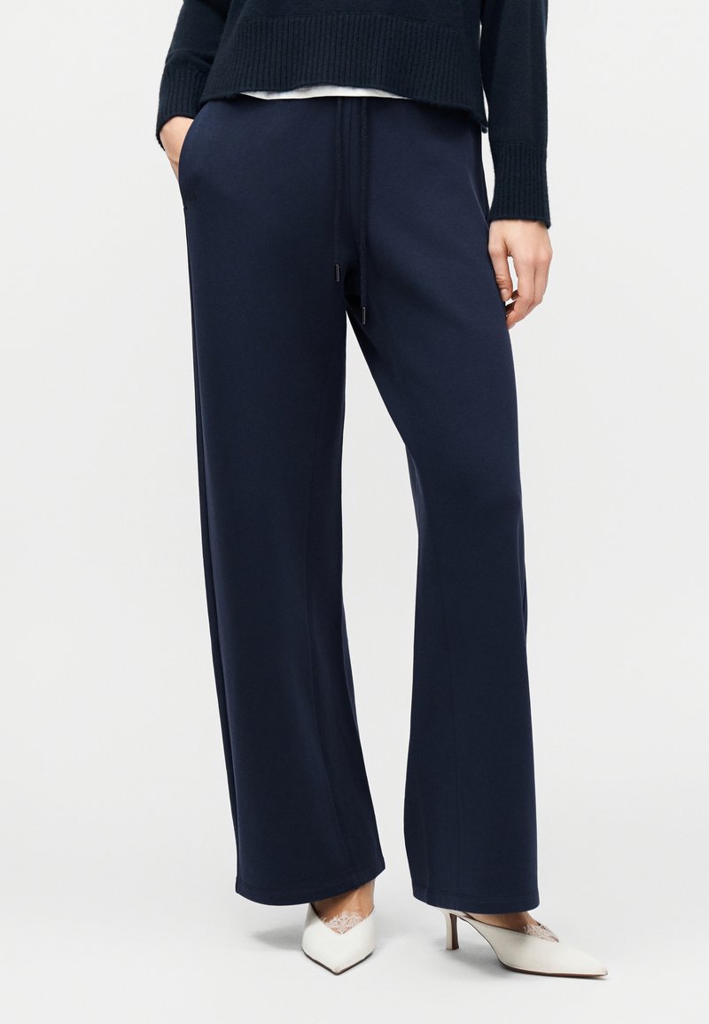 Άτομο που φοράει ναυτικό μπλε παντελόνια με φαρδύ κόψιμο και ζωνάκι με κορδόνια στη μέση, μαύρο πουλόβερ και λευκές μυτερές γόβες με δαντελένιες κάλτσες, στέκεται με τα χέρια στις τσέπες.