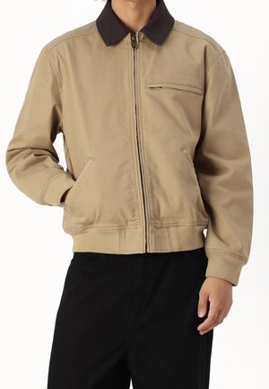 Mężczyzna ubrany w beżową kurtkę zapinaną na suwak z ciemnym kołnierzem i kieszeniami, w połączeniu z czarnymi spodniami, zgięta ręka, ręka w kieszeni.