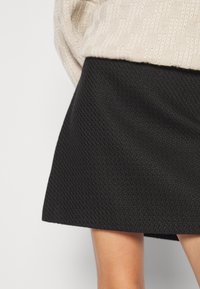 Pull beige en maille rentré dans une mini-jupe noire texturée en ligne A montrant des jambes jusqu'au genou sur un fond uni.