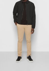 Černá lehká bomber bunda, hnědé tričko, béžové slim-fit kalhoty a černé tenisky s bílou podešví. Hladká tkanina, minimalistický design.