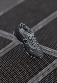 Siva sportska cipela s mrežastim i sintetičkim materijalom, s crnim akcentima, zelenim obilježjima, zaobljenim vrhom i teksturiranom potplatom.