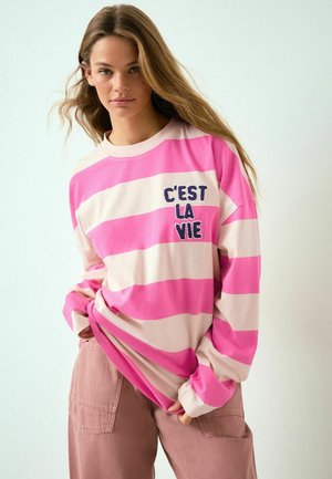 Rosa und cremefarben gestreiftes Langarmshirt mit "C’est la vie" in Marineblau auf der Brust gestickt, kombiniert mit locker sitzenden rosa Hosen.