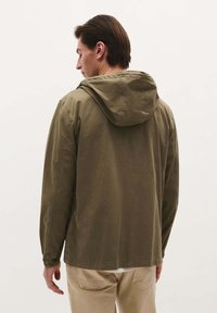 Bărbat cu spatele, purtând o jachetă cu glugă de culoare verde măslin și pantaloni bej, pe un fundal simplu, deschis la culoare.