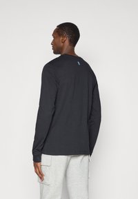 Mann trägt ein schwarzes Langarmshirt mit einem kleinen blauen NBA-Logo im oberen Rückenbereich, kombiniert mit hellgrauen Cargo-Hosen.