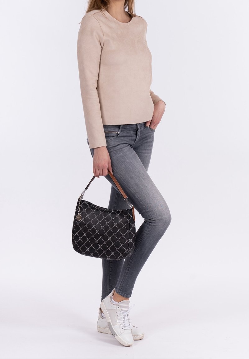 Femme portant un haut à manches longues beige et un jean gris, tenant un sac à main noir à motif avec une poignée marron, baskets blanches sur fond blanc.