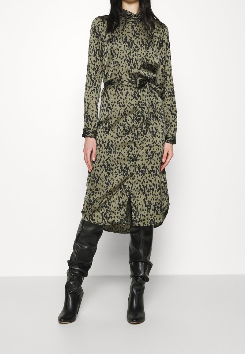 Femme portant une robe longue à manches longues vert olive avec des taches abstraites noires, taille ceinturée, et des bottes en cuir noires montant jusqu'aux genoux.