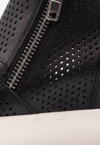 Zapato de cuero negro perforado con detalle de cremallera plateada, textura suave, suela de goma blanca y diseño abierto para ventilación.