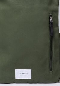 Section d'un sac en tissu vert montrant une fermeture éclair noire verticale avec un tirette et une étiquette rectangulaire blanche portant le texte "SANDQVIST".