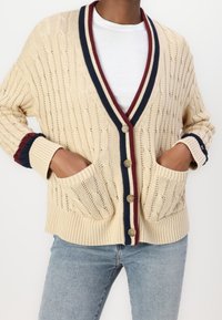 Cardigan beige en tricot avec motif torsadé, bordures côtelées bleu marine et bordeaux, poches avant, et fermeture à cinq boutons.