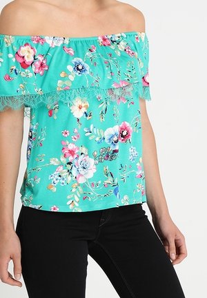 Mujer vestida con blusa floral turquesa con hombros descubiertos y ribete de encaje, combinada con pantalones ajustados negros, posando frente a un fondo liso.