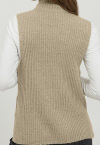 Beige värillinen ribbineuleliivi, jossa korkea kaulus, pehmeä pinta ja sivuhalkiot. Päällä valkoinen pitkähihaisen paita. Yksinkertainen, toiminnallinen muotoilu.