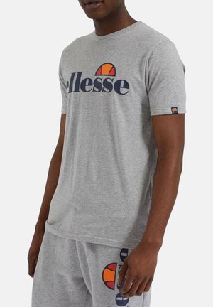 Mężczyzna w szarej koszulce Ellesse i pasujących spodenkach, z wyrazistym logo Ellesse nadrukowanym na koszulce i spodenkach.