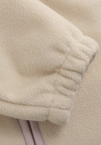 Cremefarbener Fleece-Stoff mit strukturierter Oberfläche. Verfügt über ein elastisches Bündchen und einen sichtbaren lila Reißverschlussakzent. Weiches und leichtes Design.