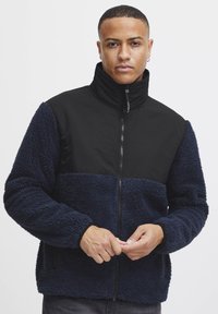 Fleece jacket featuring a navy and black color block design, zip closure, high collar, and textured fabric with side pockets.