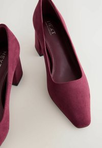 Zapatos de tacón alto de ante burdeos con punta cuadrada y tacón ancho, que presentan una textura suave y un diseño minimalista.