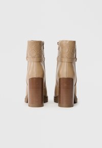 Bottes ankle beiges en matériau synthétique, talons empilés marron, tige texturée avec un motif de logo embossé et fermetures à glissière sur les côtés.