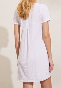 Femme blonde portant une robe blanche à manches courtes avec un pli au dos et une bordure en dentelle à l'ourlet, posant devant un fond beige.