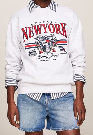 Osoba ubrana w jasnoszarą bluzę z czerwonym napisem i emblematem „New York”, pod którą znajduje się koszula w paski z kołnierzykiem, oraz niebieskie szorty z denimu.