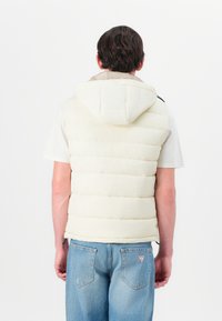 Helle beige Daunenweste mit Kapuze, versehen mit horizontalen Steppnähten und einer weichen Textur. Getragen über einem grauen T-Shirt und blauen Jeans.