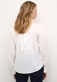 Fehér, hosszú ujjú blúz, enyhe fényességgel, puffadt vállakkal és ívelt szegéllyel. Az anyag könnyűnek és simának tűnik.
