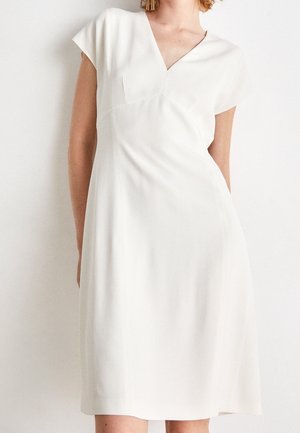 Femme portant une robe blanche longueur genou avec manches courtes et encolure en V, debout devant un fond blanc uni.