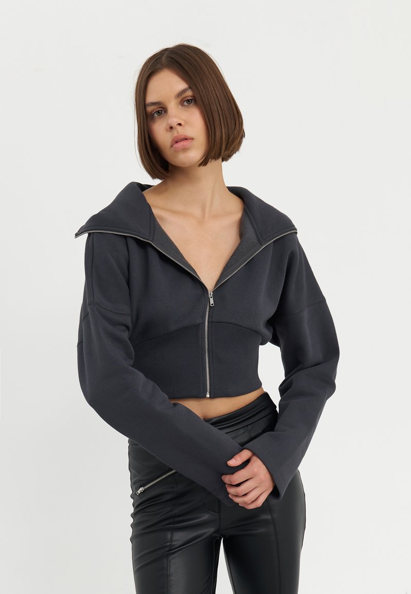 Skraćena zip-up dukserica tamnosive boje, s velikim ovratnikom, rebrastom teksturom i glatkim patentnim zatvaračem. Uparen s crnim uskim hlačama.