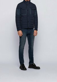 Námorná péřová bunda s vysokým límcem, dvěma náprsními kapsami a zapínáním na knoflíky. V kombinaci s tmavými džíny a černými šněrovacími botami.