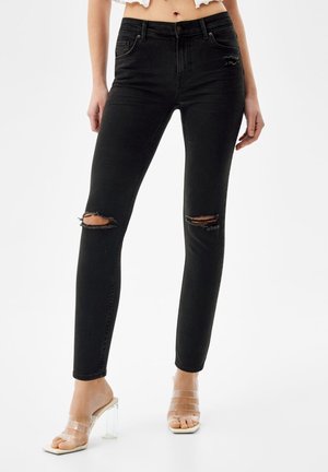 Femme portant un jean skinny noir taille haute avec des déchirures aux genoux, associé à des sandales à talons hauts à brides transparentes sur un fond uni.