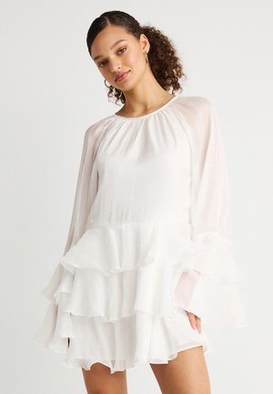 Kobieta o kręconych włosach, ubrana w białą sukienkę z długim rękawem z warstwowymi falbanami i kolczykami kołami, stojąca na jednolitym tle.