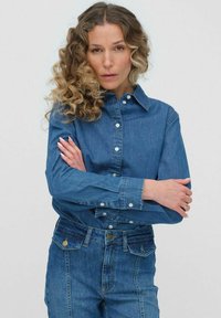 Koszula dżinsowa w średnim niebieskim kolorze, z klasycznym kołnierzykiem, zapięciem na guziki z przodu oraz zapinanymi mankietami. Posiada regularny krój. Noszona z dobrze dopasowanymi niebieskimi dżinsami.