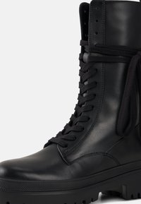 Czarny skórzany but z wysoką cholewką, okrągłym noskiem, grubą podeszwą i płaskimi sznurowadłami. Gładka faktura z wyraźnym przeszyciem i oczkami.