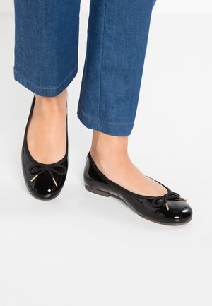 Voeten met zwarte lakleren ballerina's met strikjes, gecombineerd met blauwe cropped broeken op een witte ondergrond.