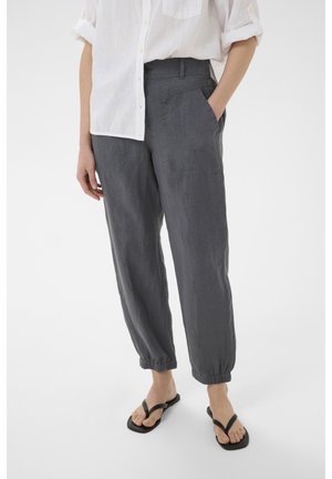 Persona que lleva pantalones sueltos de color gris oscuro con puños elásticos, una camisa blanca abotonada con las mangas remangadas y chanclas negras, de pie.