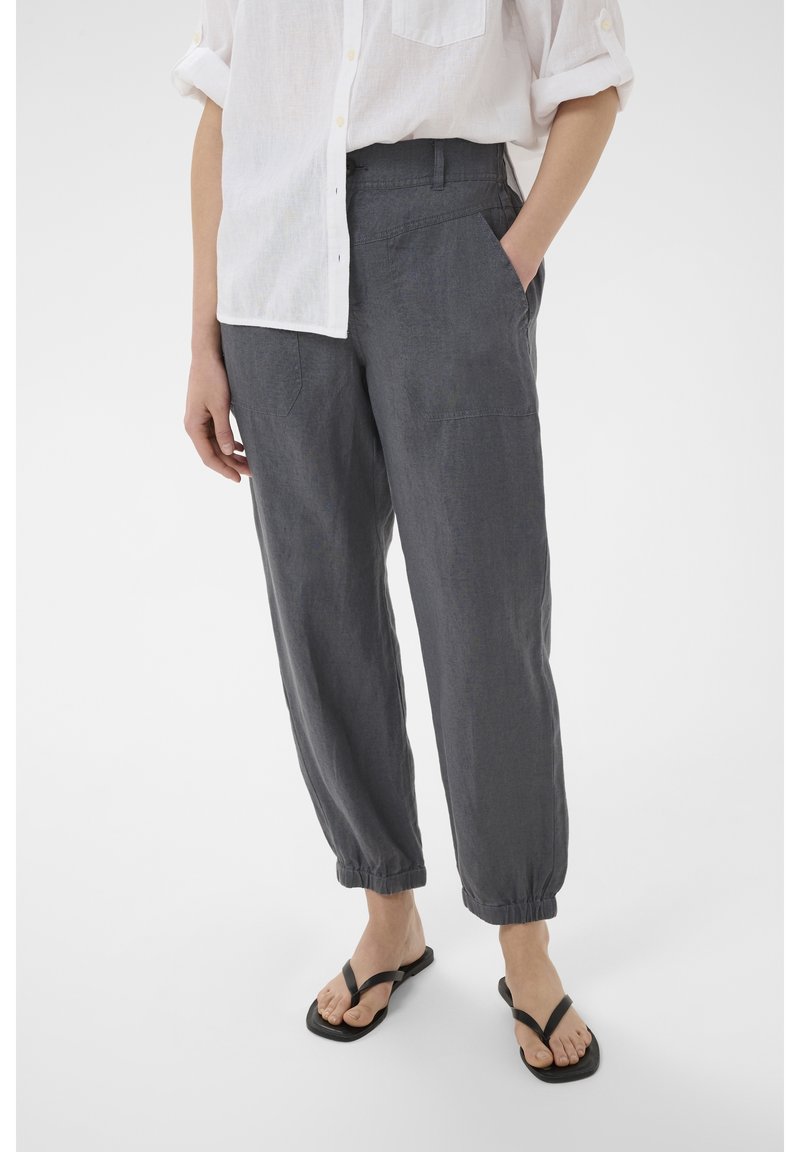 Personne portant un pantalon ample gris foncé avec des poignets élastiques, une chemise blanche boutonnée avec les manches retroussées, et des tongs noires, debout.
