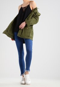 Olijfgroene parka jack, zwart veter-topje, blauwe skinny jeans met rafelrand, en witte sneakers. Casual gelaagd outfit.
