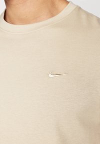 Šviesiai ruda medvilnės marškinėliai su apvaliu iškirpimu, su mažu, siuvinėtu sidabro spalvos Nike logotipu kairėje krūtinės pusėje. Lygus paviršius.
