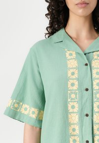 Groen shirt met korte mouwen, voorzien van een knopenloop aan de voorkant, met gele bloempatronenaccenten langs de knopenlijst en de mouwen. Zachte textuur.