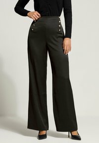Hoge taille zwarte wijde broek van soepel materiaal, met gouden knoopaccenten aan de voorkant. Gecombineerd met puntige zwarte hakken.