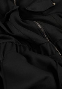 Vêtement noir avec une fermeture éclair visible et une texture de tissu froncé. Comprend des poches plates et un matériau doux et lisse.