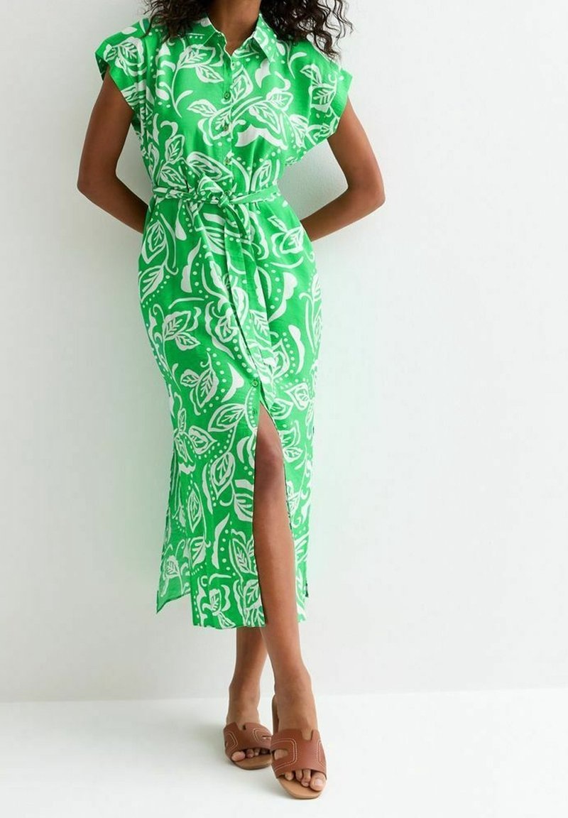 Vihreä kukallinen midi-mekko, jossa on lyhyet hihat, napitus edessä ja itsesidottava vyö; sisältää reiteen saakka ulottuvan halkion ja sileän kankaan pinnan. Ruskeat liukuvarvassandaalit.