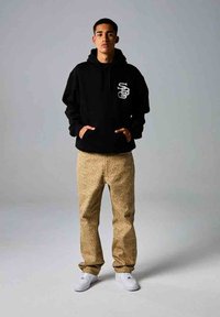 Sudadera negra con capucha y logotipo en el pecho, combinada con pantalones beige texturizados con un diseño estampado. Modelo lleva zapatillas blancas.